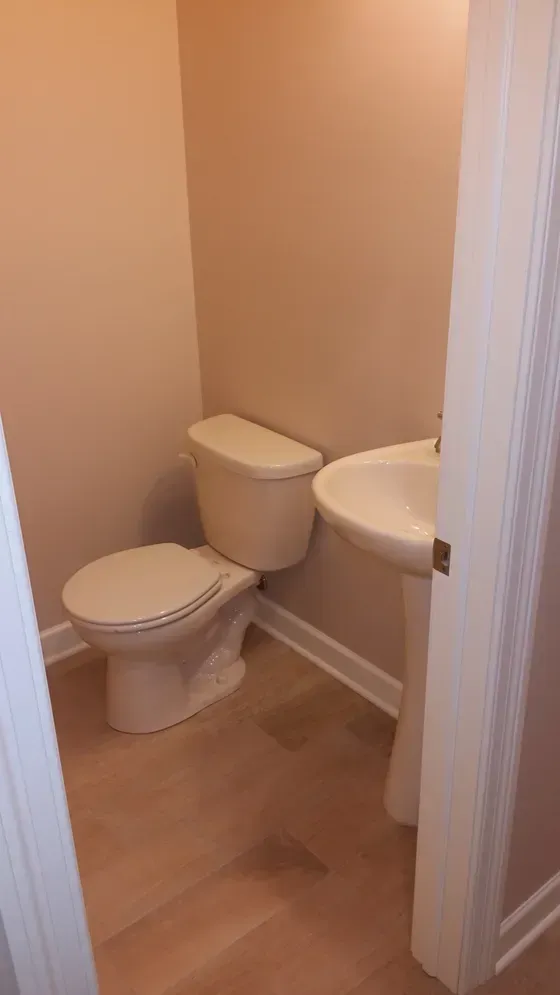 A small bathroom with a toilet, sink, and tan walls and flooring, viewed from the doorway.