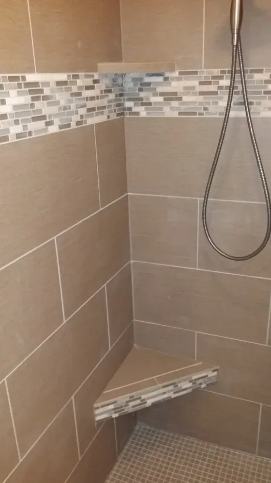 Shower with tan tiles, mosaic accent, triangular corner seat, and shelf.
