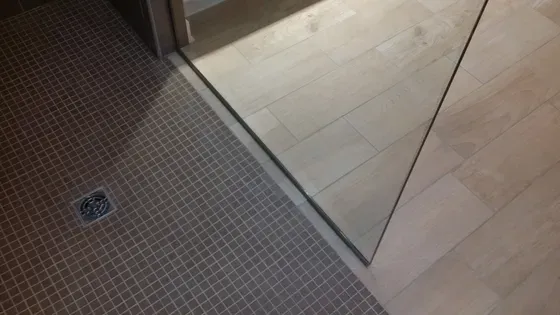 Shower with textured brown tile floor, light wood floor, glass shower door, and drain.