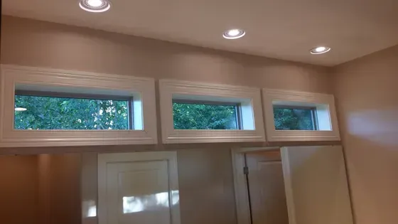 Three small rectangular windows in white trim, above closed white doors, with recessed ceiling lights, in a room with beige walls.