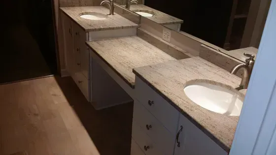Bathroom vanity with granite countertop and two sinks. Light cabinets, mirrored wall, and wood flooring.