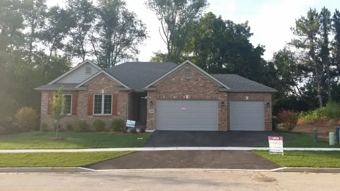 Brick ranch house with two-car garage, gray doors, and a For Sale sign on the lawn.