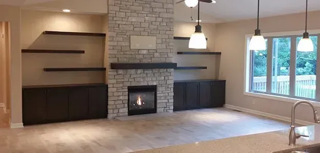 Living room with fireplace, stone wall, floating shelves, dark cabinets, and window.