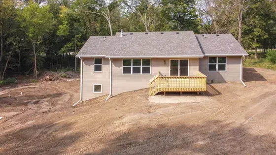 A single-story beige house with a wooden deck and a gray roof, set in a cleared area surrounded by trees.