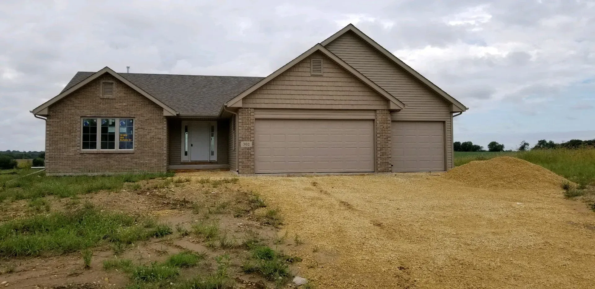 New house with light brown siding and garage doors, gravel driveway, and grassy yard under cloudy sky.