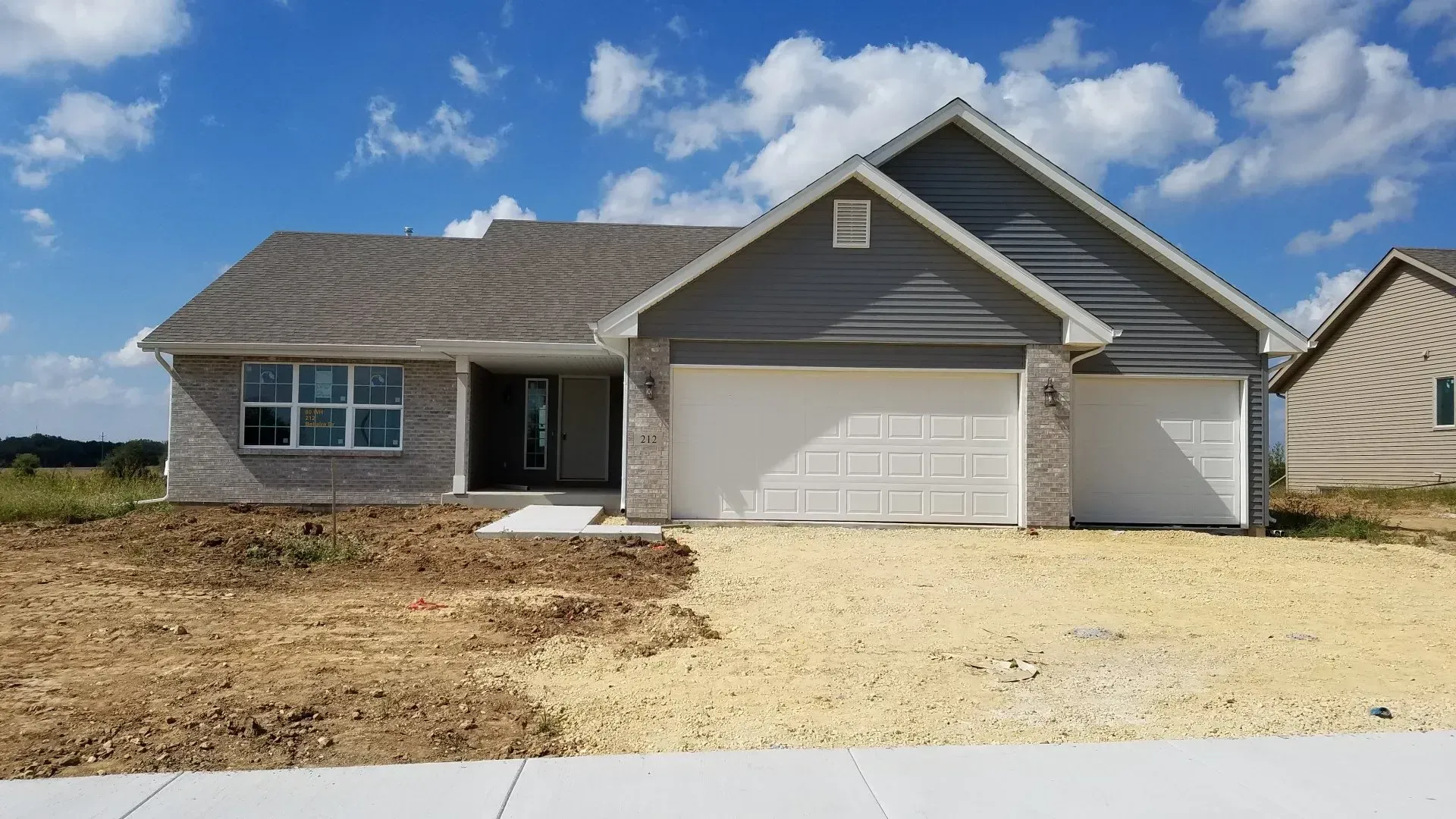 Single-story house with gray siding and garage, light brick, and dirt yard on a sunny day.