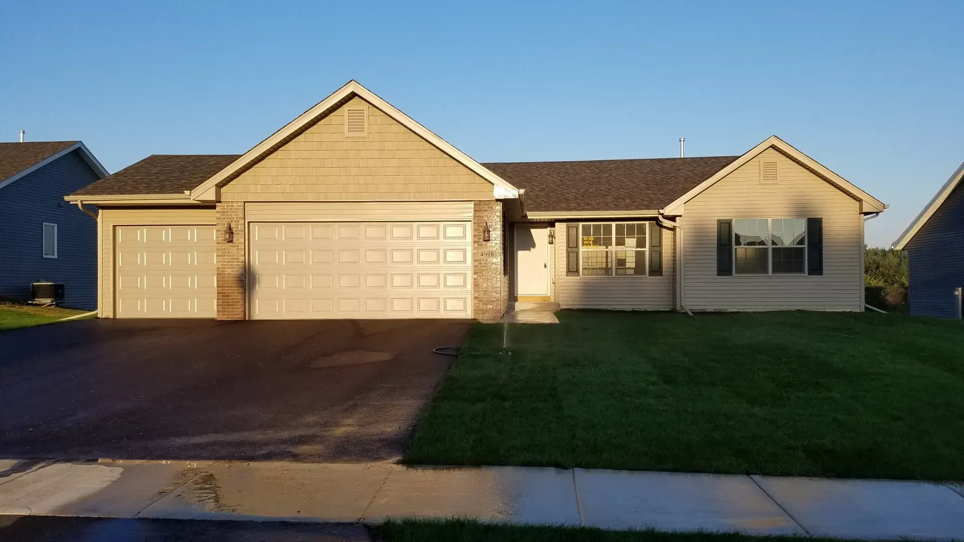 Ranch-style house with a tan facade and brown roof, a two-car garage, and green lawn on a sunny day.