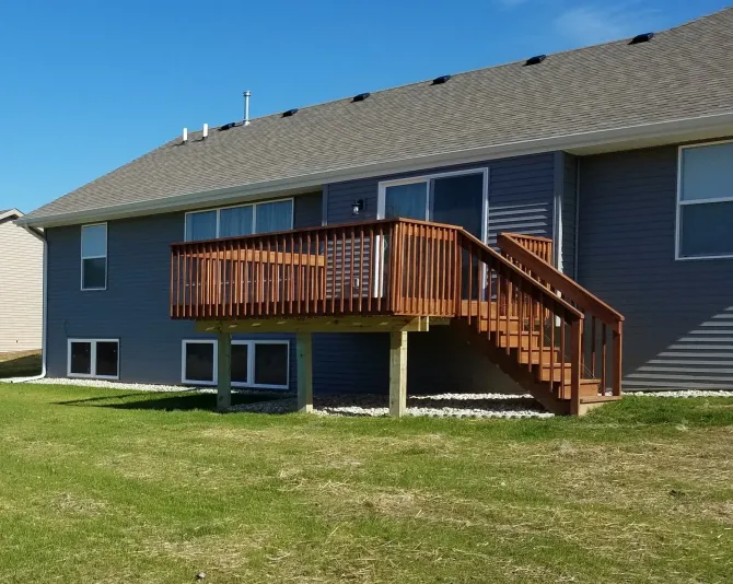 Wooden deck with stairs, attached to a gray house, on a grassy lawn under a blue sky.