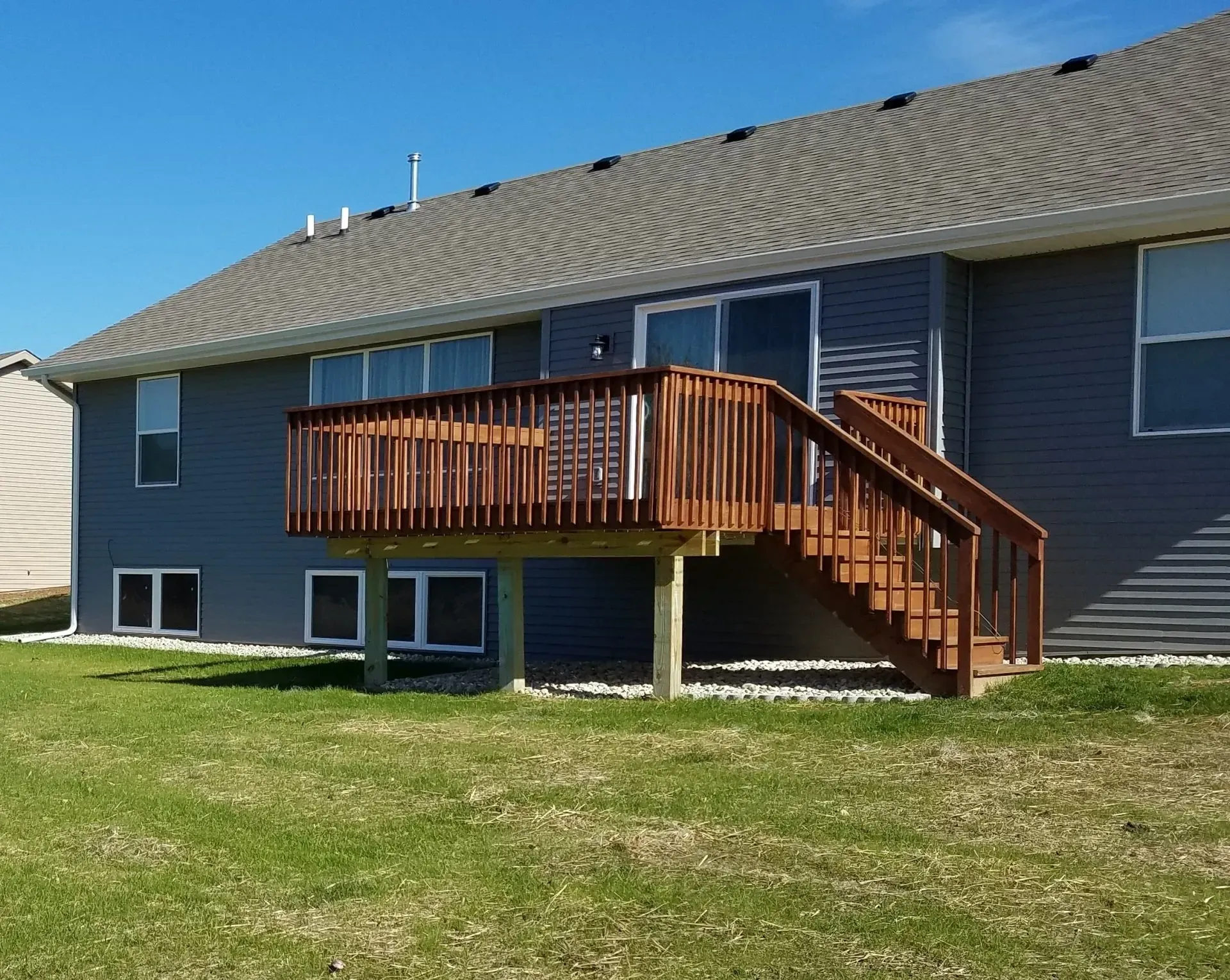 Wooden deck with stairs attached to a house with blue siding. Bright green grass and a clear blue sky.