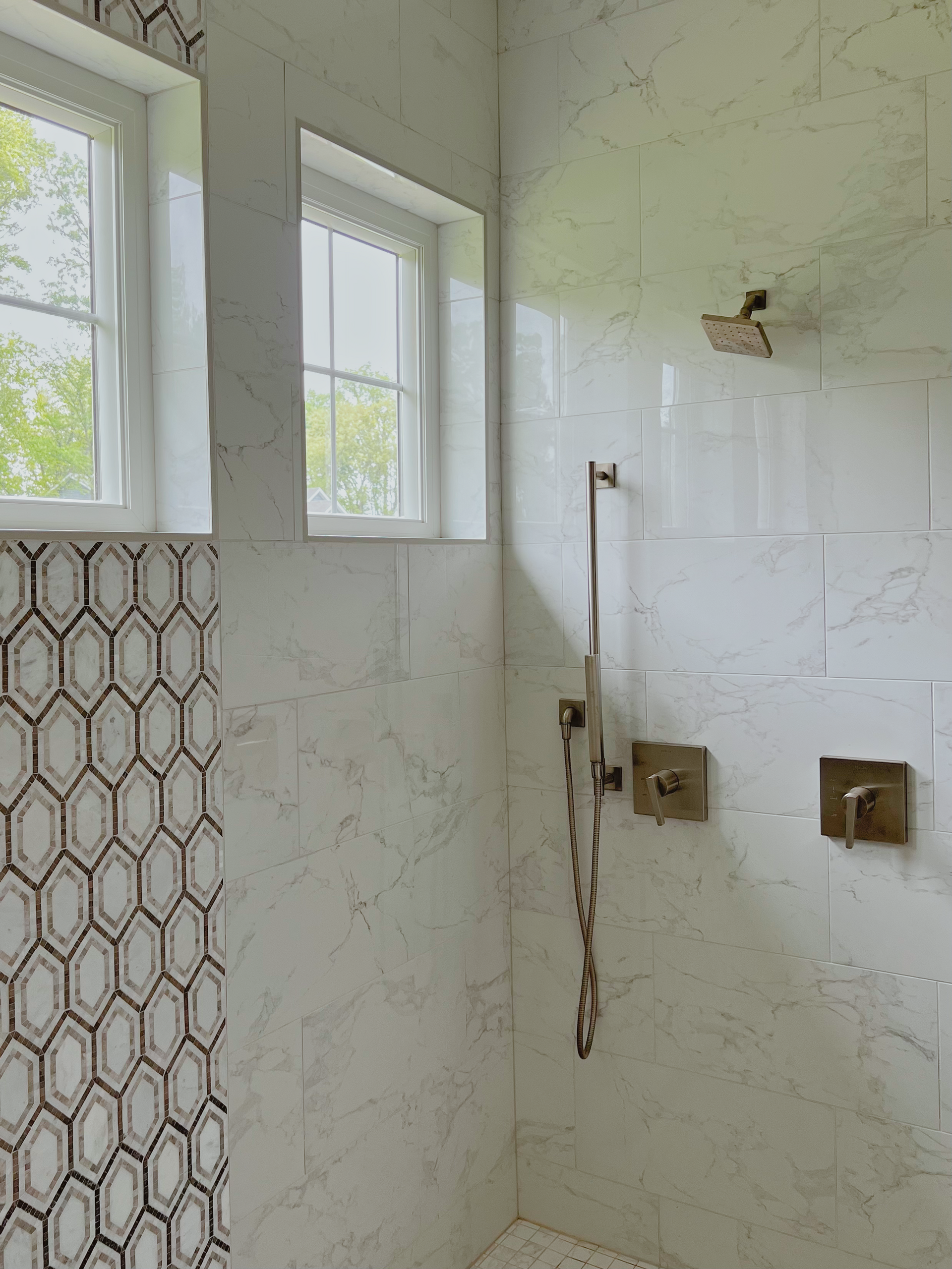 Bathroom with marble-look tile walls and a hexagon accent wall. Gold shower fixtures. Two windows at top.