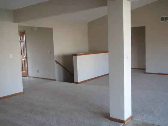 Empty living room with tan walls, carpet, and a staircase. A white column is in the foreground.