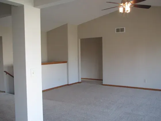 Empty living room with beige walls, tan carpet, and a high ceiling with a ceiling fan.