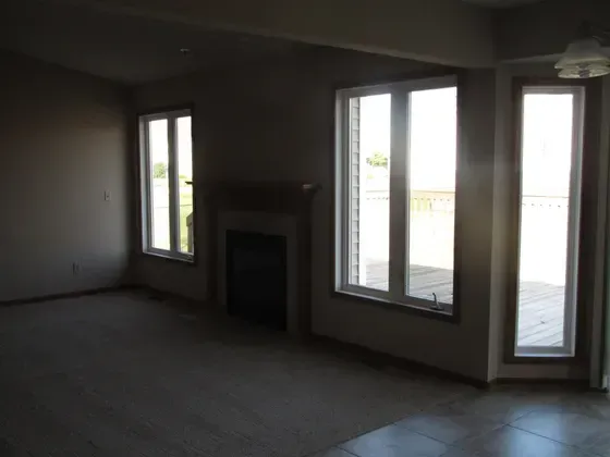 Empty living room with fireplace and large windows, neutral walls, light-colored carpet.