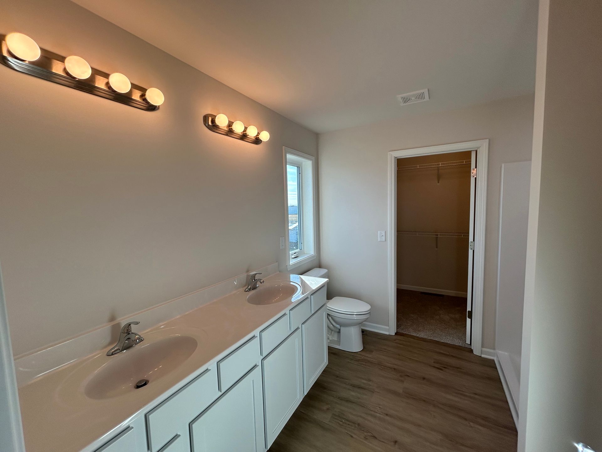 Bathroom with double sinks, toilet, and walk-in closet. Light wood-look flooring and neutral walls.