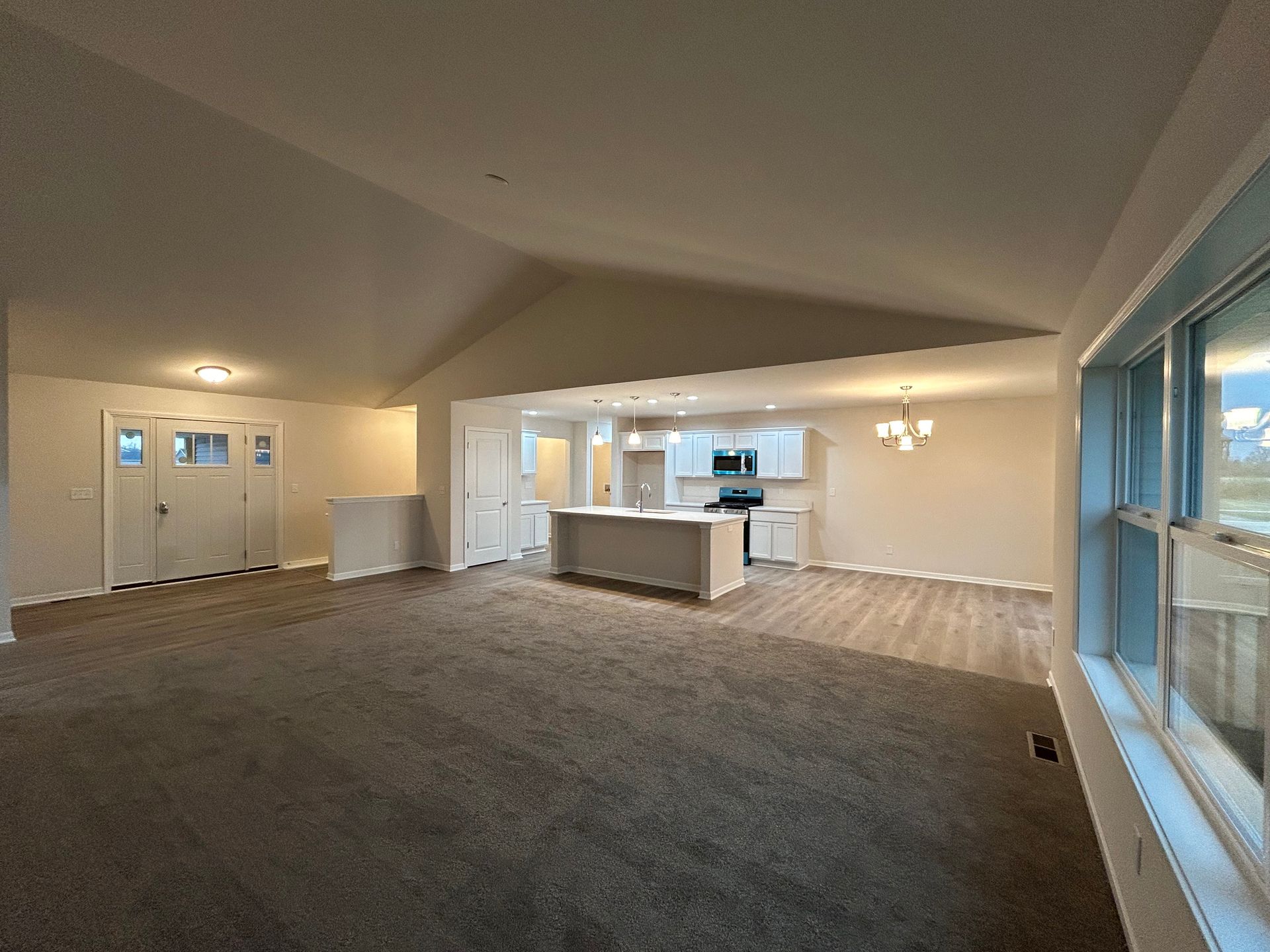 Open-concept living space with vaulted ceiling, kitchen, and front door. Neutral tones, gray carpet, and large window.
