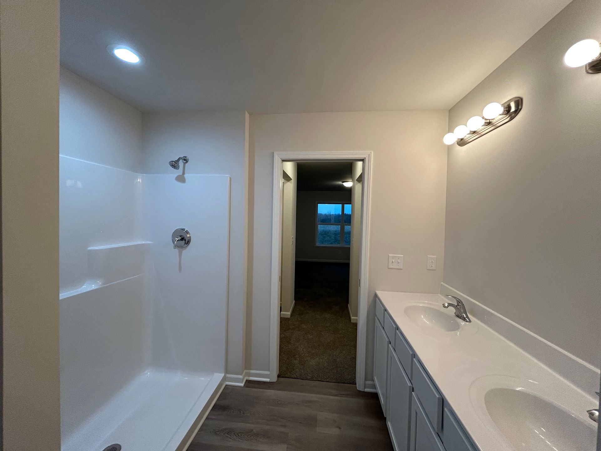 Bathroom with shower, double sink vanity, and doorway to another room. Gray, white, and wood-look flooring.