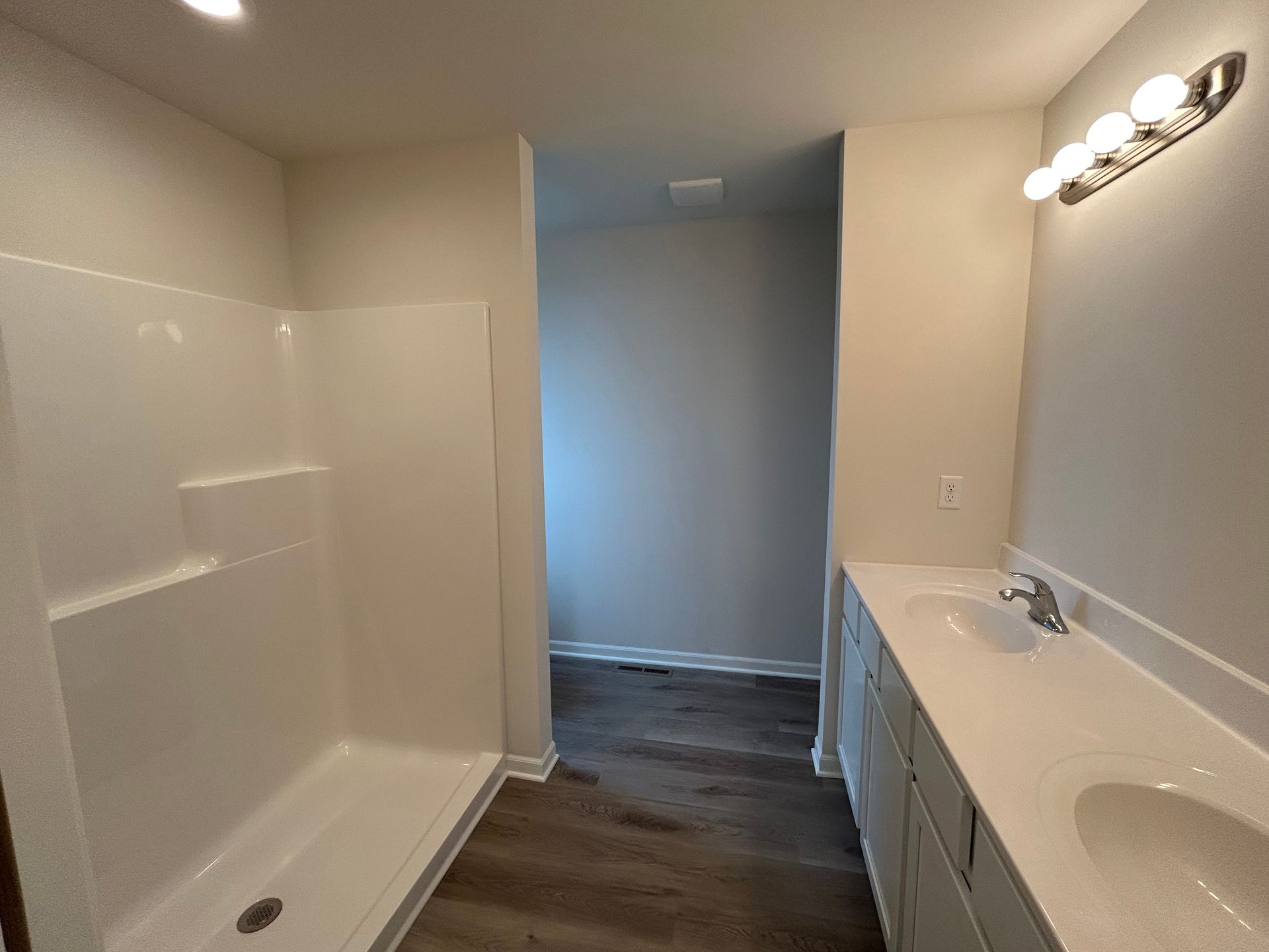 Bathroom with white shower, vanity with two sinks, and hallway with dark floor.