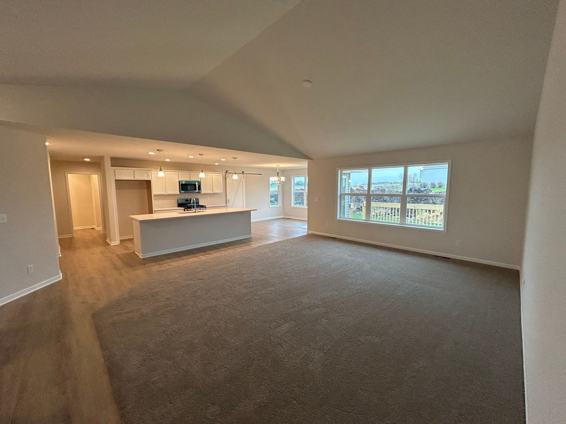 Empty living room with carpet and open kitchen. White walls and cabinetry, natural light from windows.
