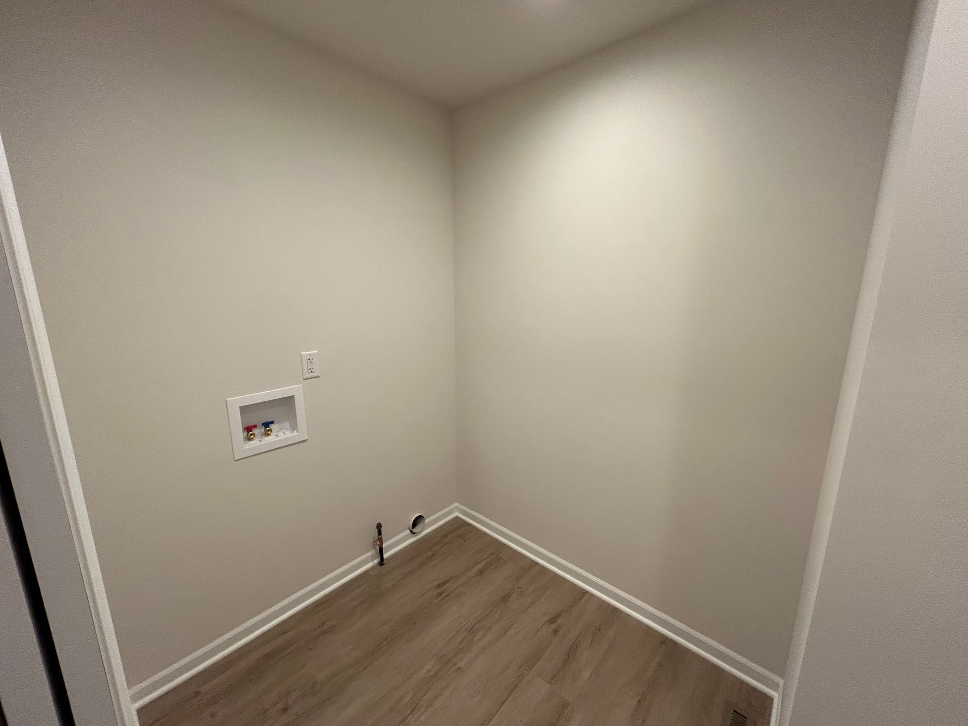 Empty laundry room with beige walls, wood-look floor, and white trim. Plumbing and electrical outlets are visible.