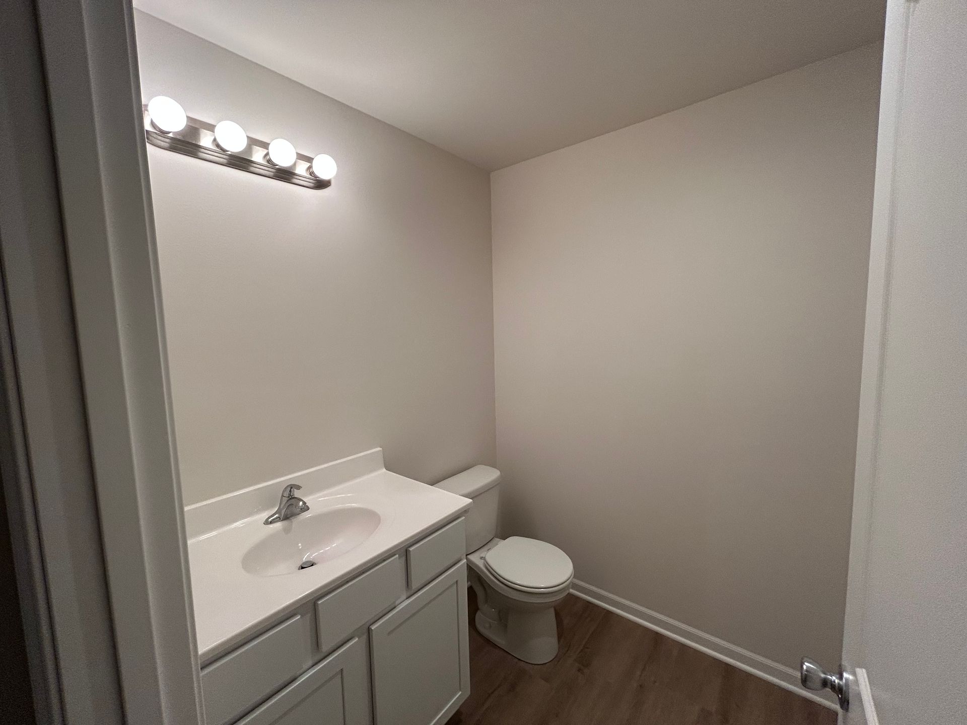 Bathroom with a vanity, toilet, and a light fixture; neutral walls, wood-look floor.