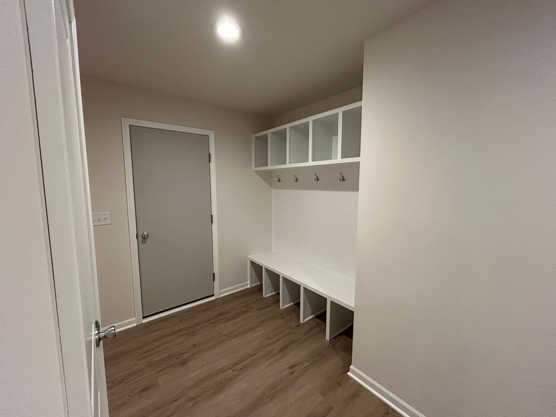 Mudroom with white built-in storage, bench, and door. Wood-look flooring and white walls.