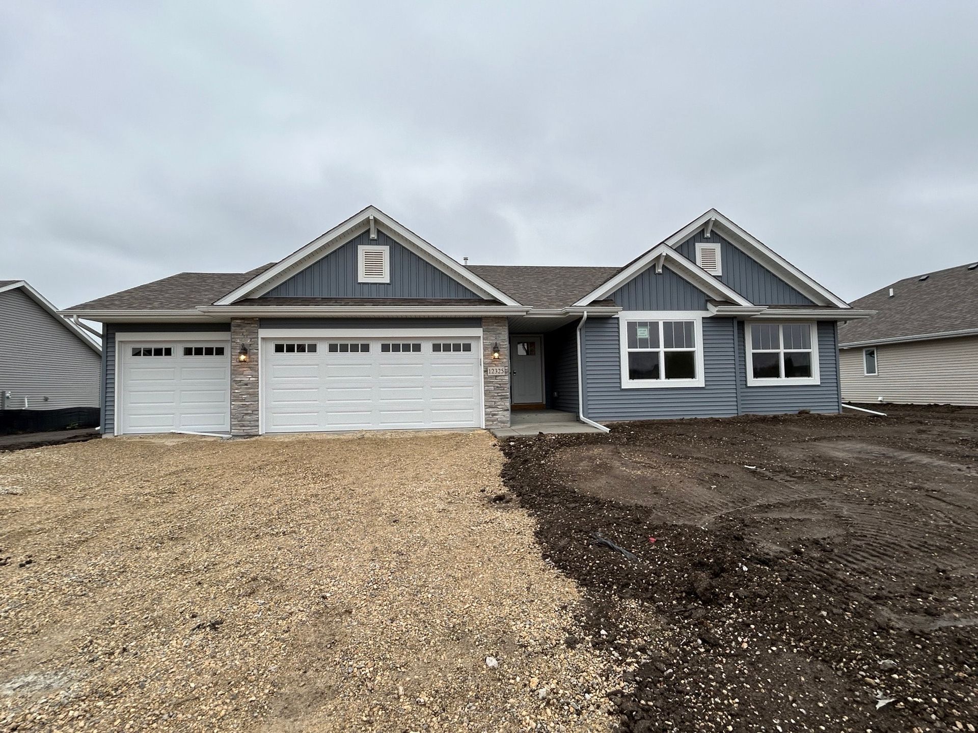 Blue and white suburban house with a three-car garage under a cloudy sky, with gravel and dirt yard.