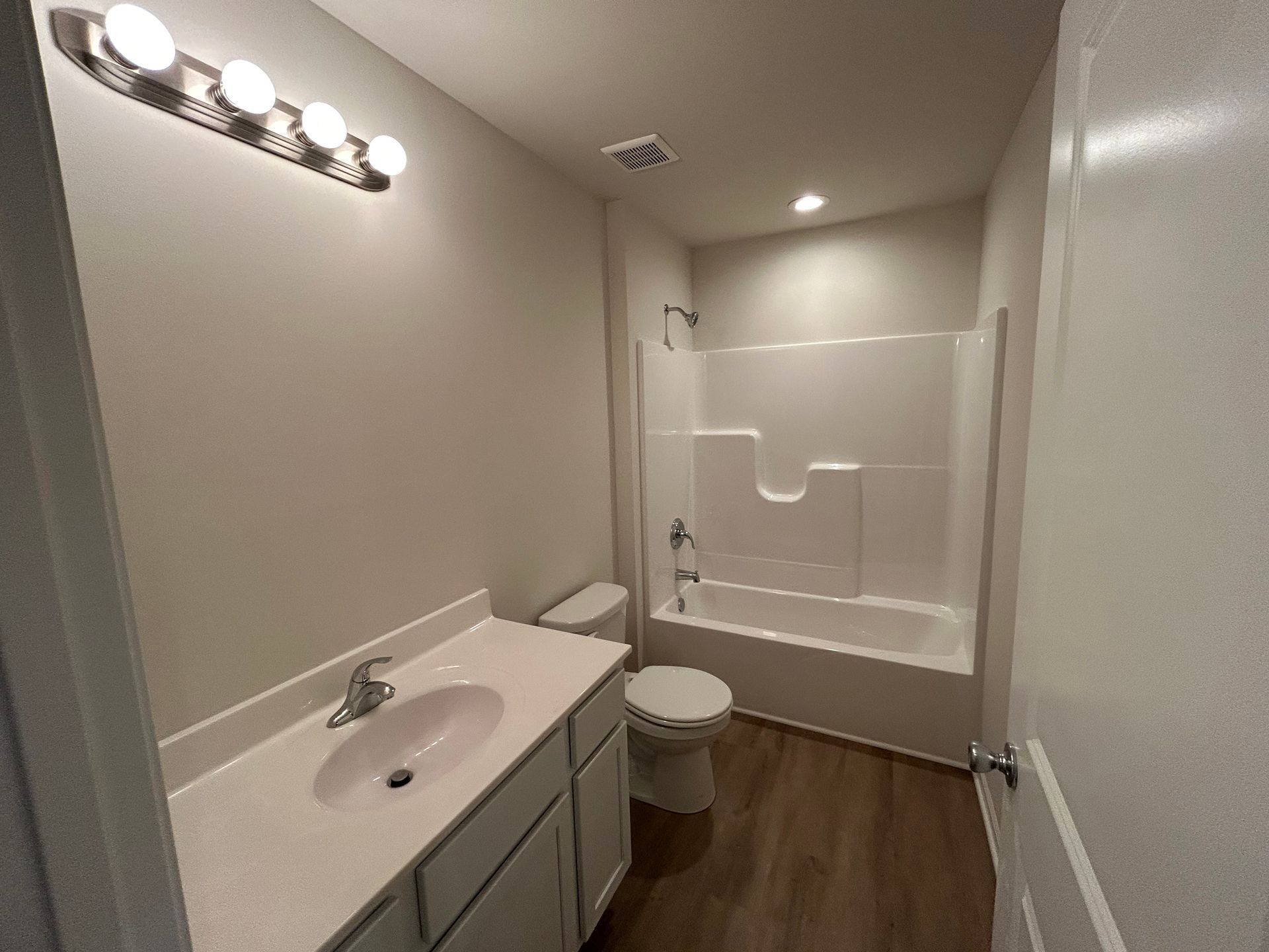 Bathroom with white tub/shower, toilet, vanity, and a row of lights. Brown flooring.