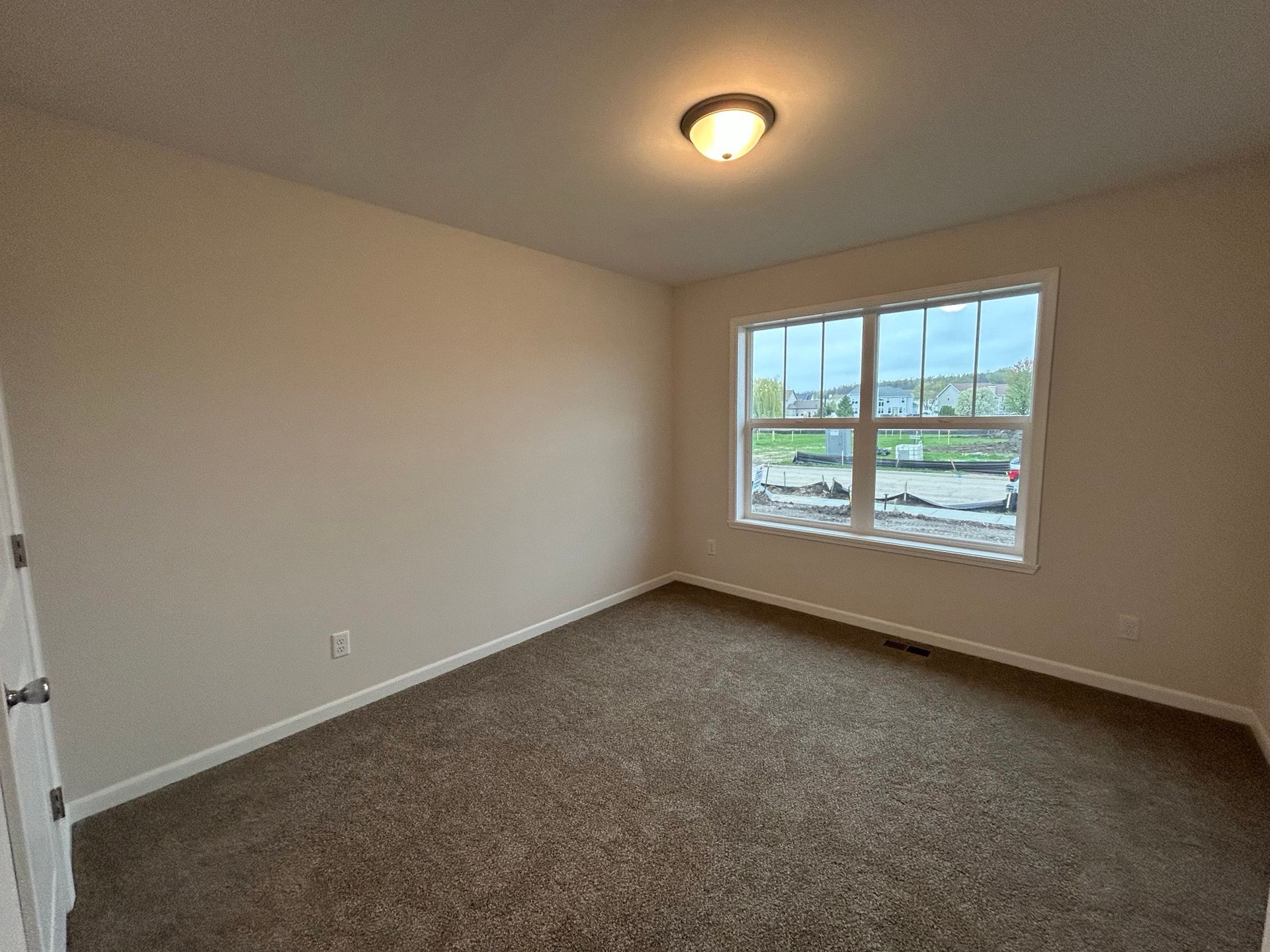 Empty bedroom with tan walls, brown carpet, and a window overlooking a construction site.