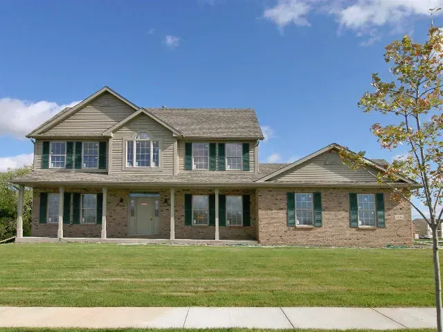 Two-story house with a brick facade, green shutters, and a large front lawn under a blue sky.