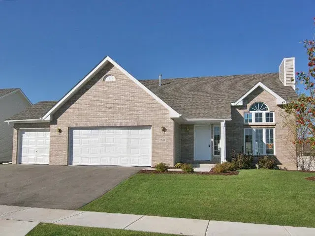 Beige brick house with a two-car garage, green lawn, and blue sky.