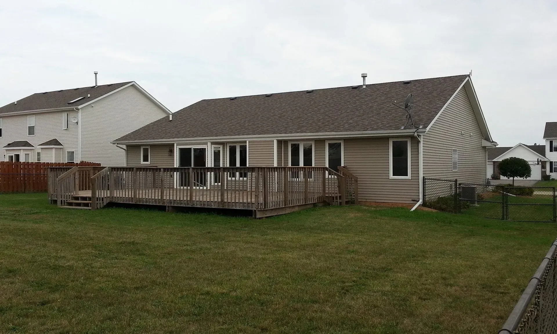 Back view of a beige house with a wooden deck, lawn, and neighboring houses on a cloudy day.