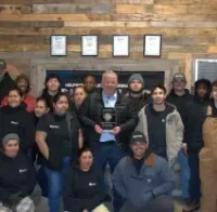 A group of people standing together indoors against a wood-paneled wall, with one person holding an award in the center.