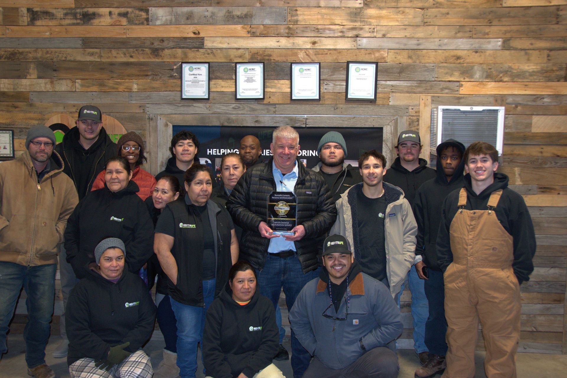 A group of people standing together indoors against a wood-paneled wall, with one person holding an award in the center.