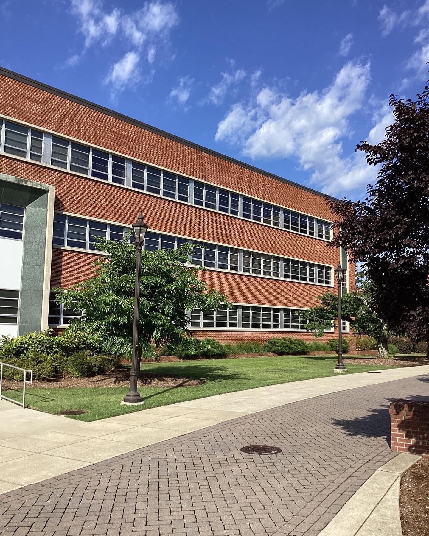 A three-story red brick building with many windows sits behind a paved walkway, green grass, and trees on a sunny day.