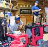 Two workers in a warehouse inspect and prepare red industrial equipment, including dehumidifiers.