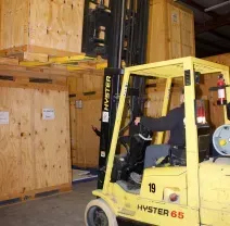 A forklift operator in a warehouse moves a large wooden shipping crate stacked on top of another.