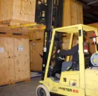 A forklift operator in a warehouse moves a large wooden shipping crate stacked on top of another.