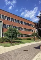 A three-story red brick building with many windows sits behind a paved walkway, green grass, and trees on a sunny day.