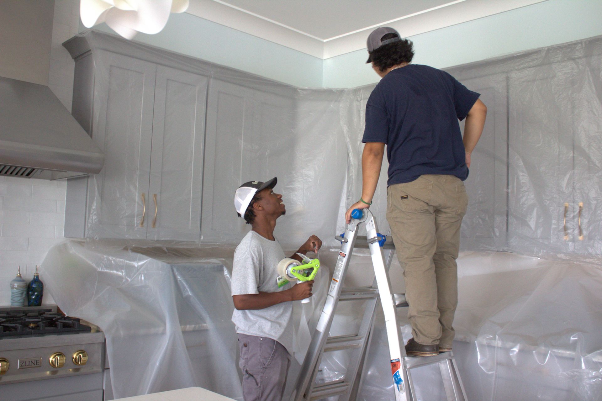 Two people preparing a kitchen for painting by covering cabinets and counters with protective plastic sheeting.