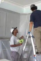 Two people preparing a kitchen for painting by covering cabinets and counters with protective plastic sheeting.