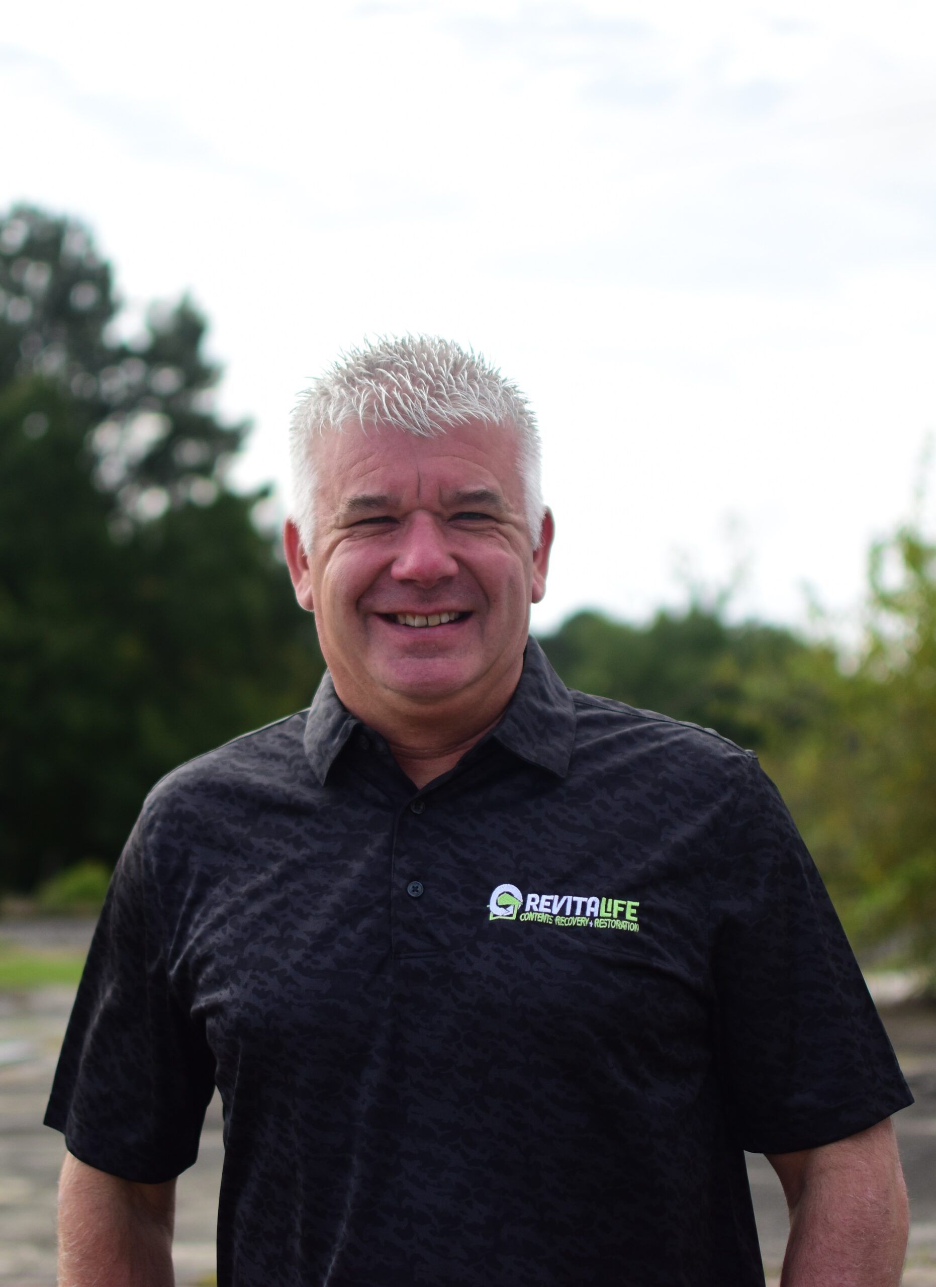 A smiling person with short light hair wearing a black polo shirt with a logo, standing outdoors in front of trees.