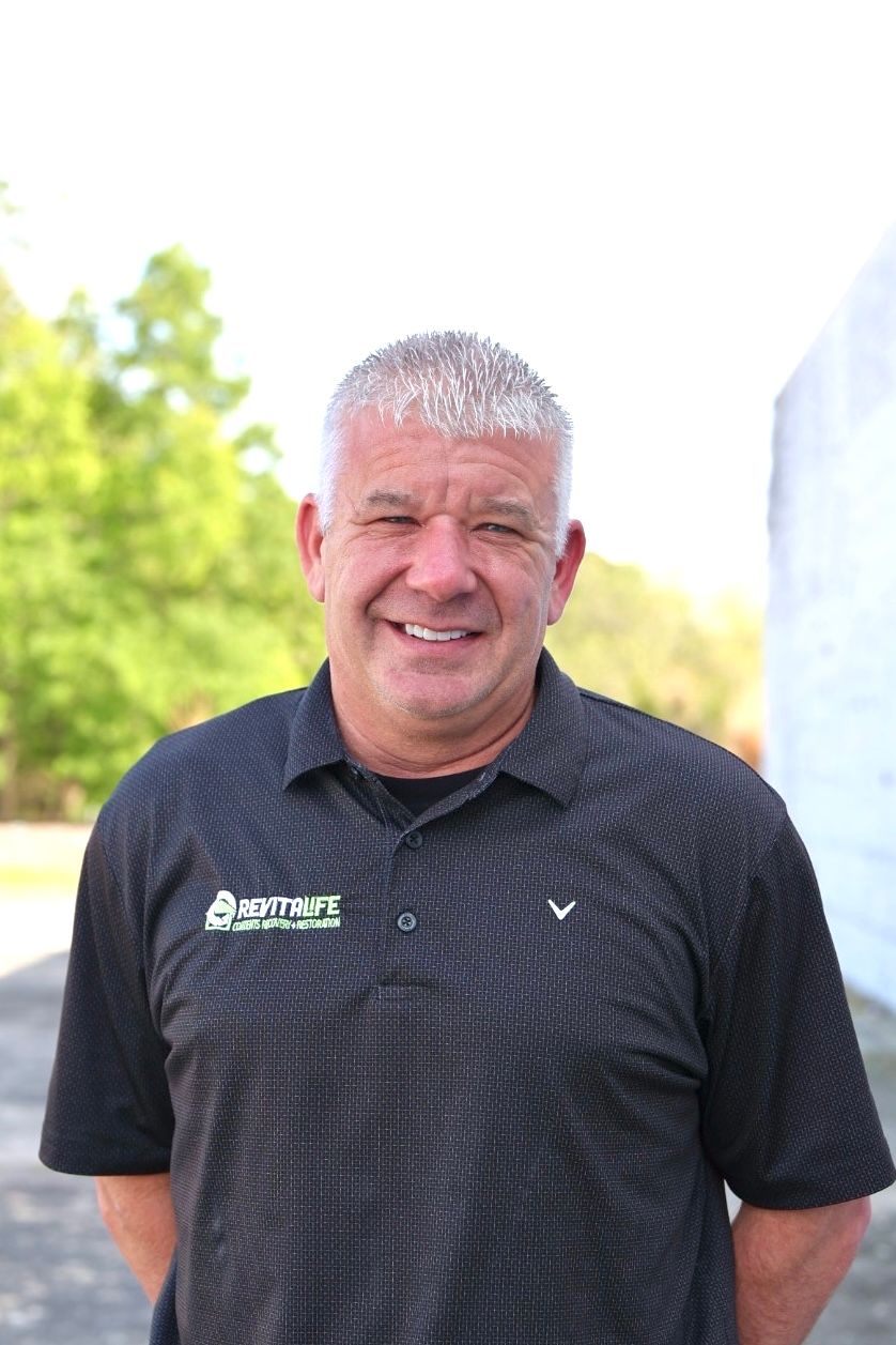A smiling person with short light hair wearing a black polo shirt with a logo, standing outdoors in front of trees.