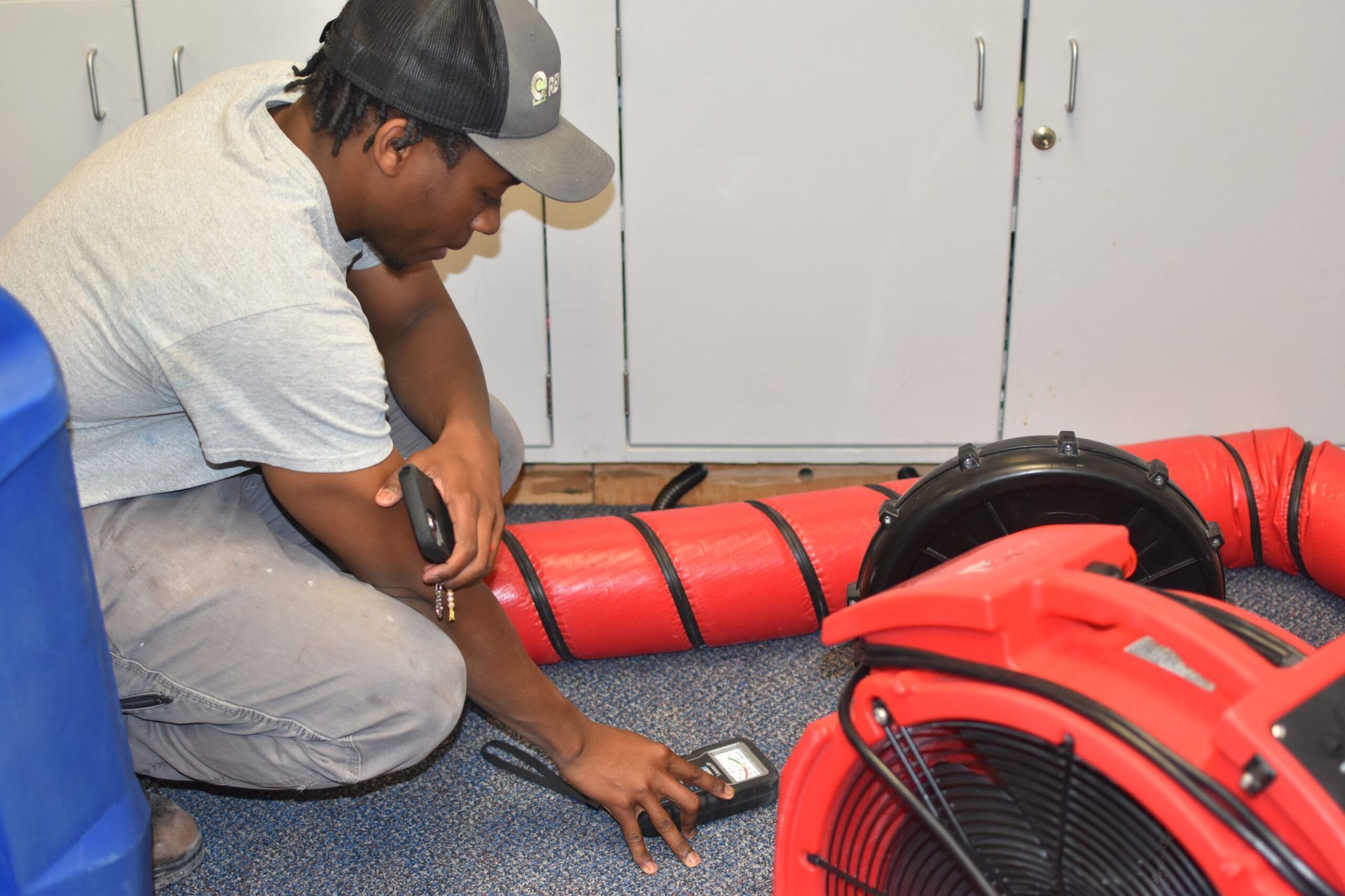 A person in a cap and gray shirt uses a handheld moisture meter on a carpet near a red industrial air mover and ducting.
