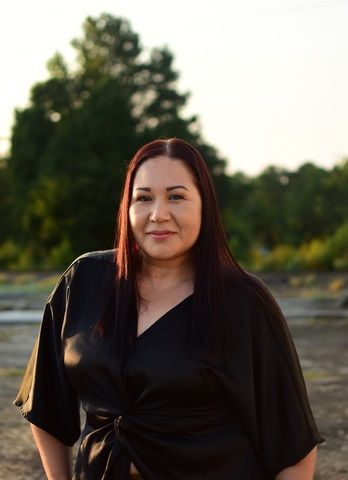 A person with dark hair wearing a black wrap top stands outdoors in front of a tree and field during sunset.