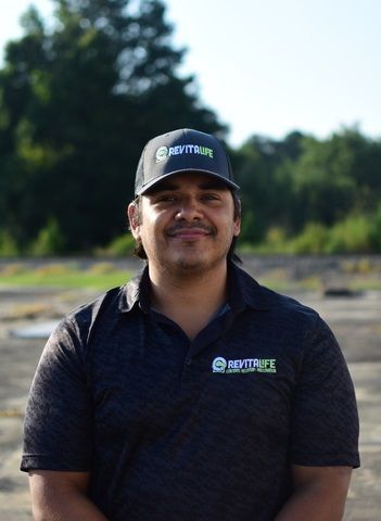 A person wearing a grey and black RevitaLife cap and polo shirt smiles while standing outdoors against a wooded backdrop.