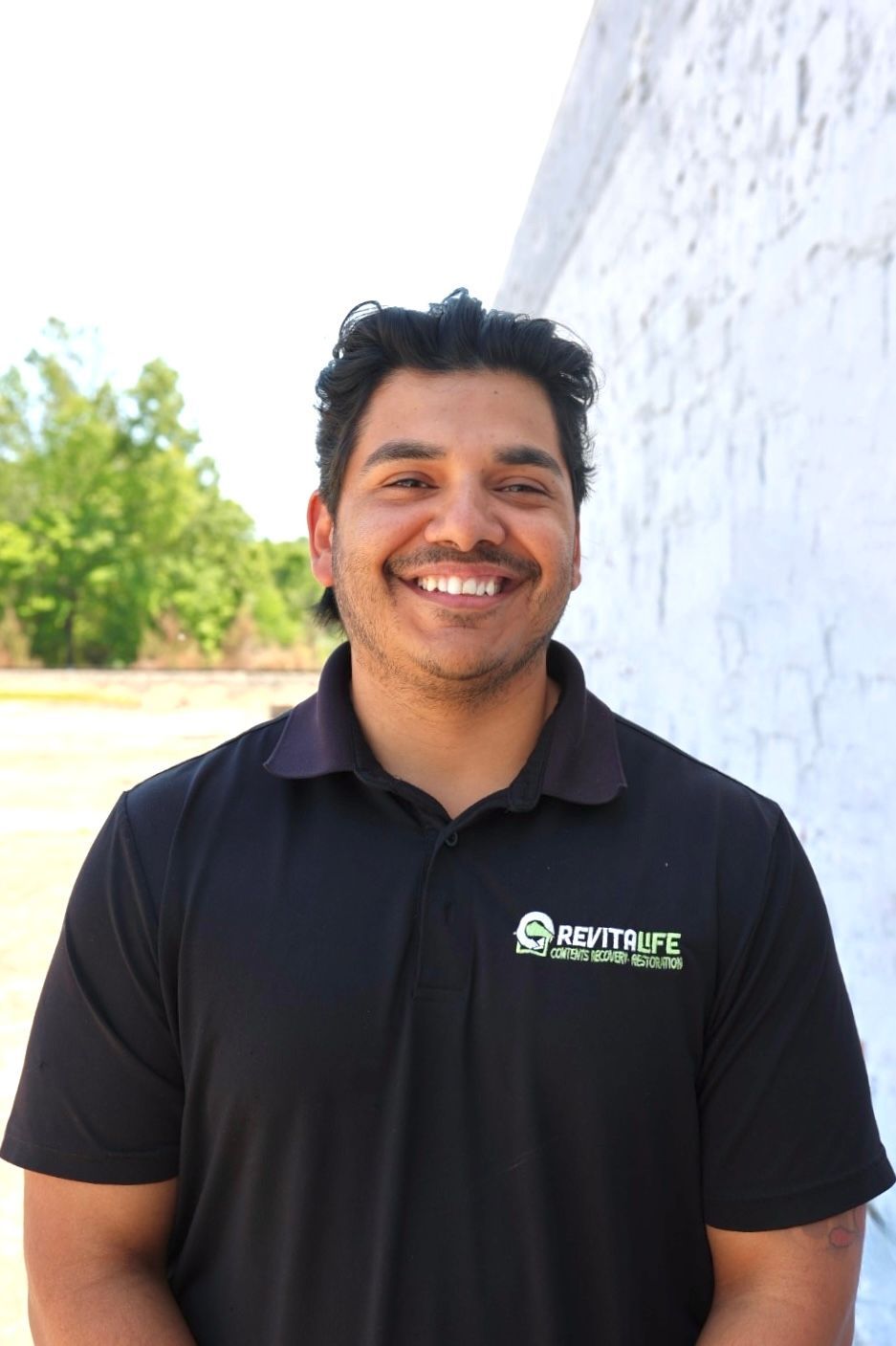 A person wearing a grey and black RevitaLife cap and polo shirt smiles while standing outdoors against a wooded backdrop.