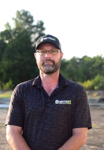 A man with a beard and glasses wears a branded black polo and cap, standing outside in a field.