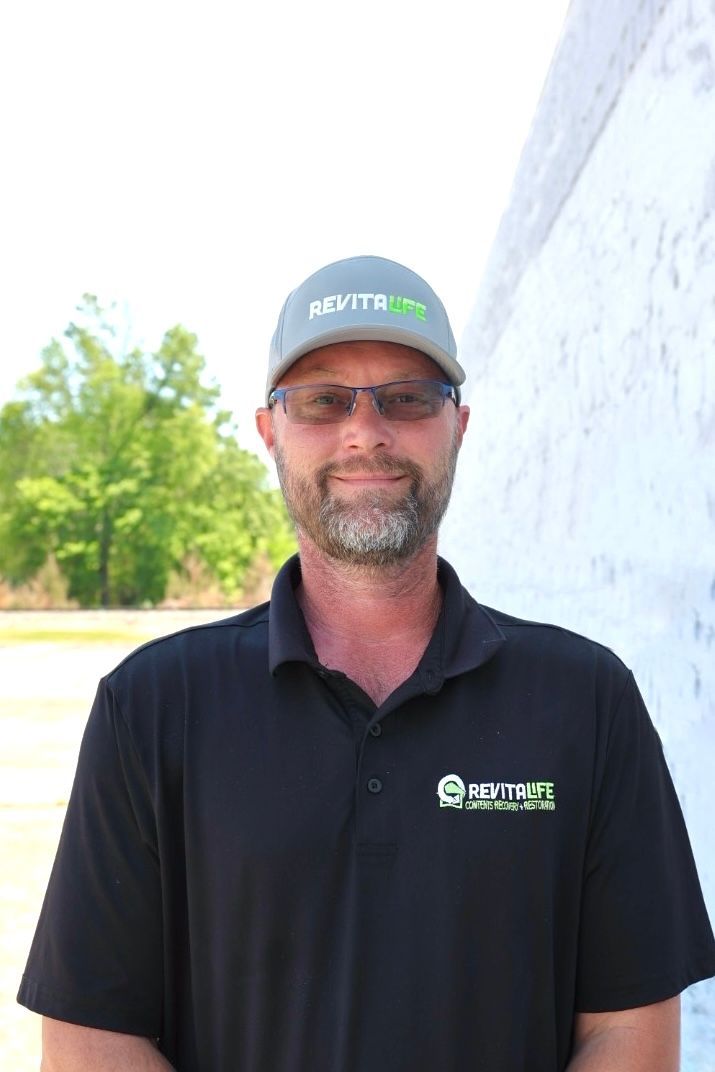 A man with a beard and glasses wears a branded black polo and cap, standing outside in a field.