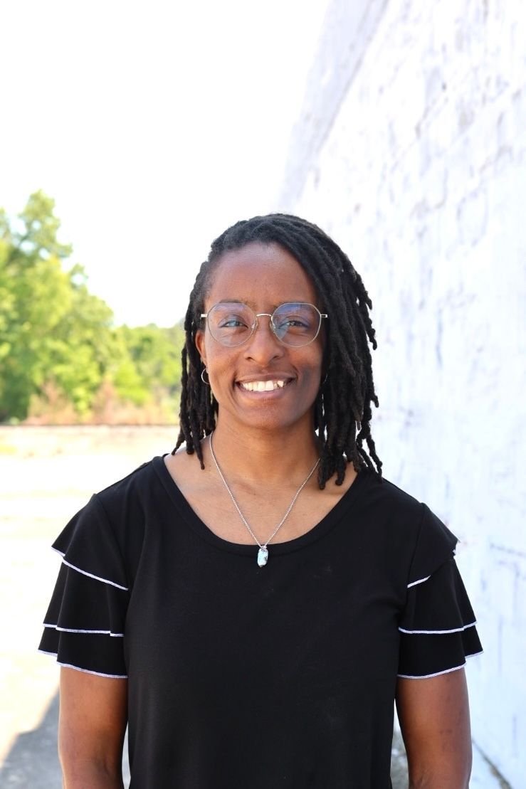 A smiling person with dark hair, wearing a black tiered-sleeve shirt and a necklace, outdoors against a tree-filled backdrop.