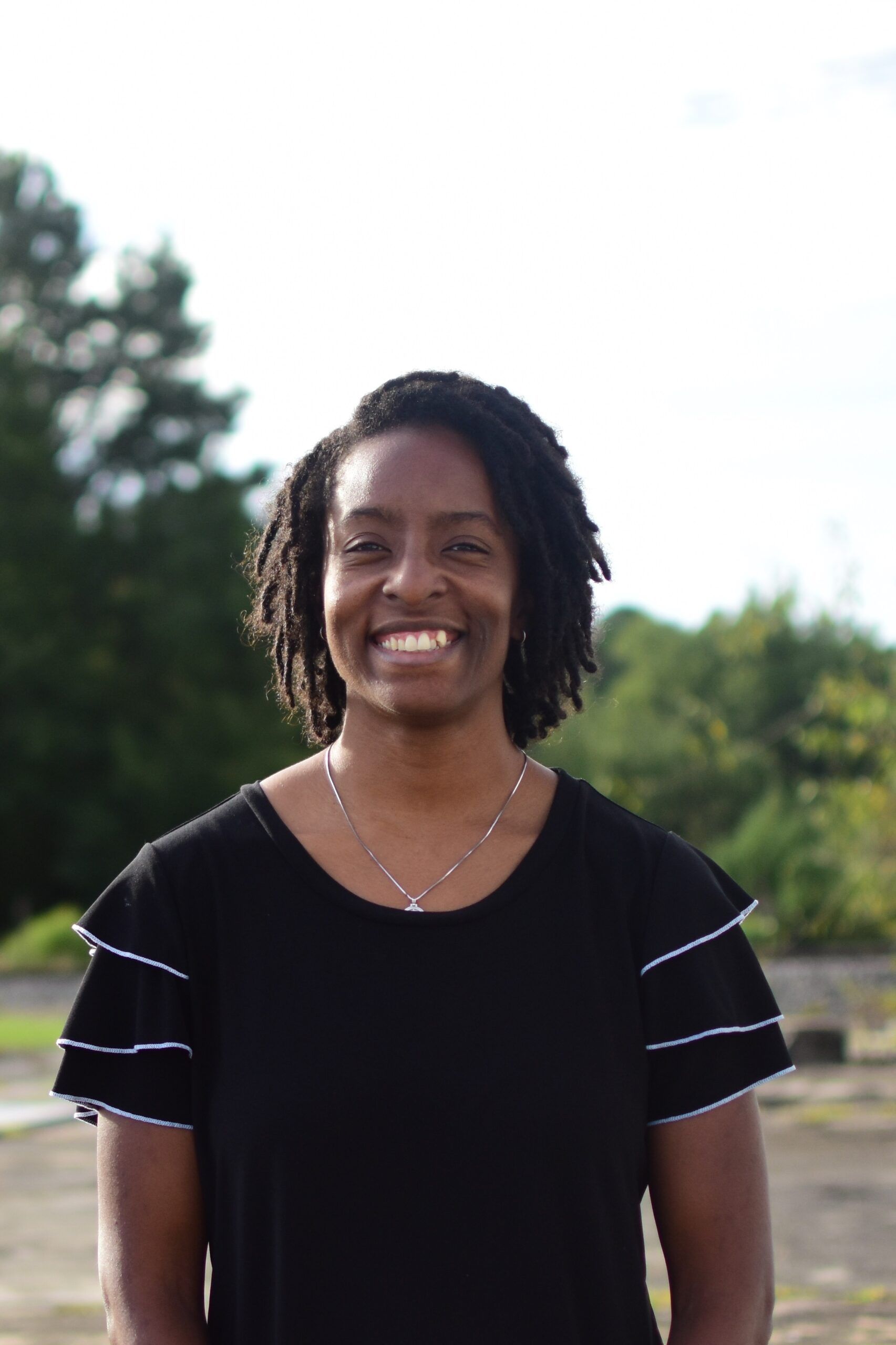 A smiling person with dark hair, wearing a black tiered-sleeve shirt and a necklace, outdoors against a tree-filled backdrop.